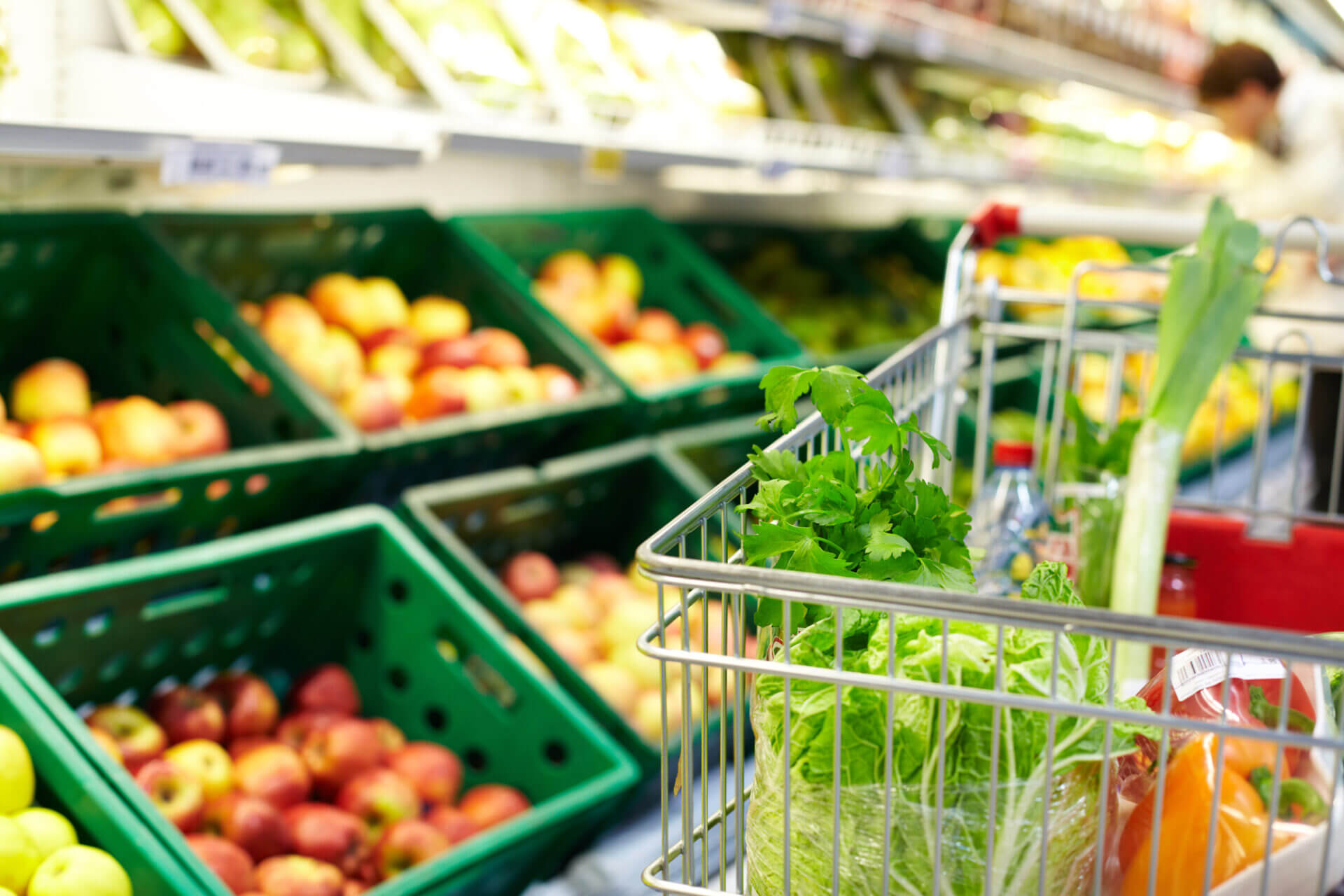 Big Climate Database, Shopping trolley filled with fresh vegetables and herbs in front of shelves of apples in a supermarket produce section.