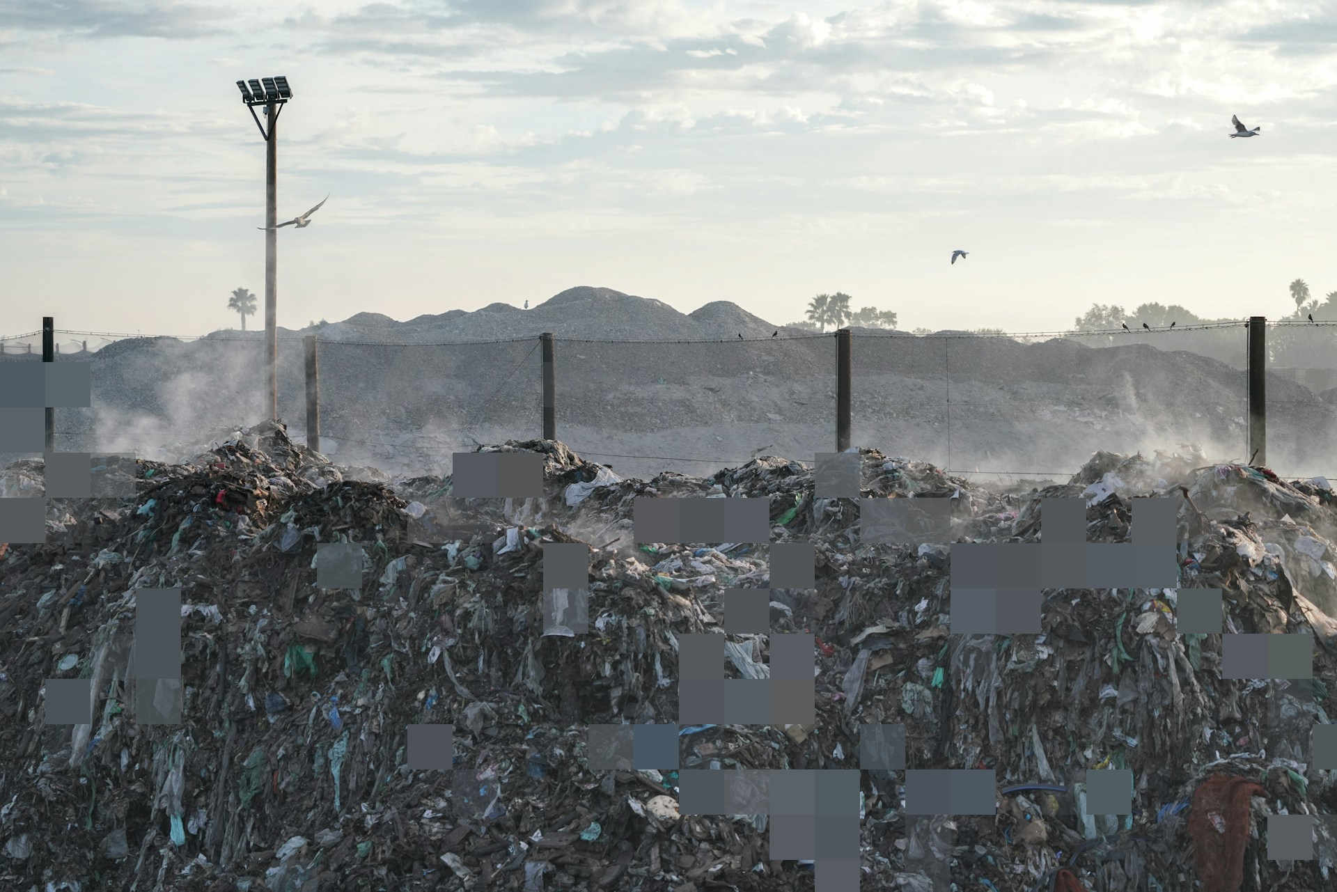 Piles of mixed waste at a landfill, with birds overhead and a selective pixel effect across the foreground.