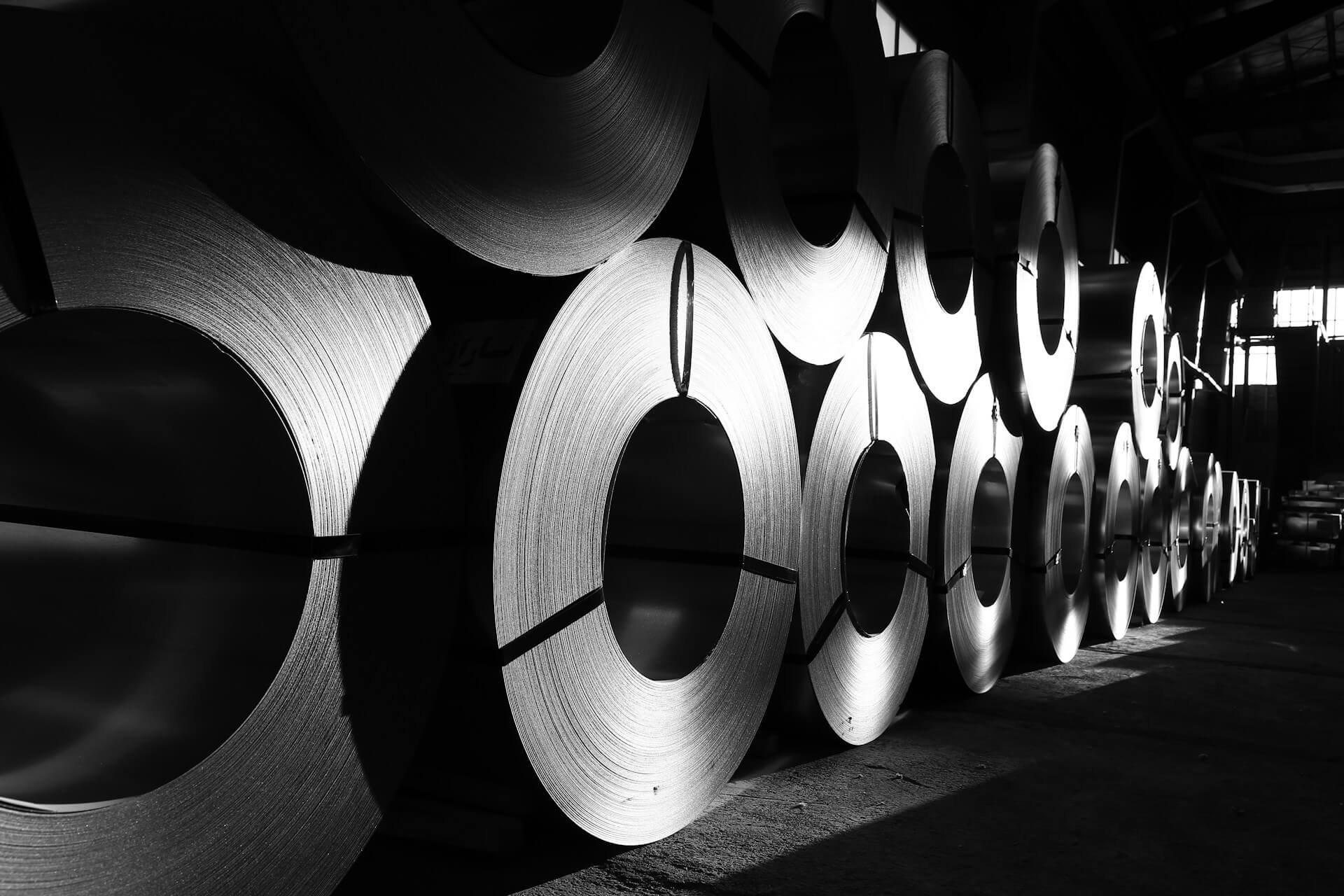 Rows of steel coils stacked inside an industrial warehouse, photographed in black and white.